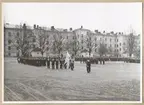 Uniformsklädda flottister marscherar över Sparres kaserngård. Ledet leds av en militär som håller i en svensk flagga. I bakgrunden syns kasern Sparre.