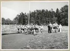 En grupp idrottsmän tävlar i löpning mot varandra längs en motionsbana. Vid sidan av står en grupp människor och ser på. I bakgrunden syns en skog.