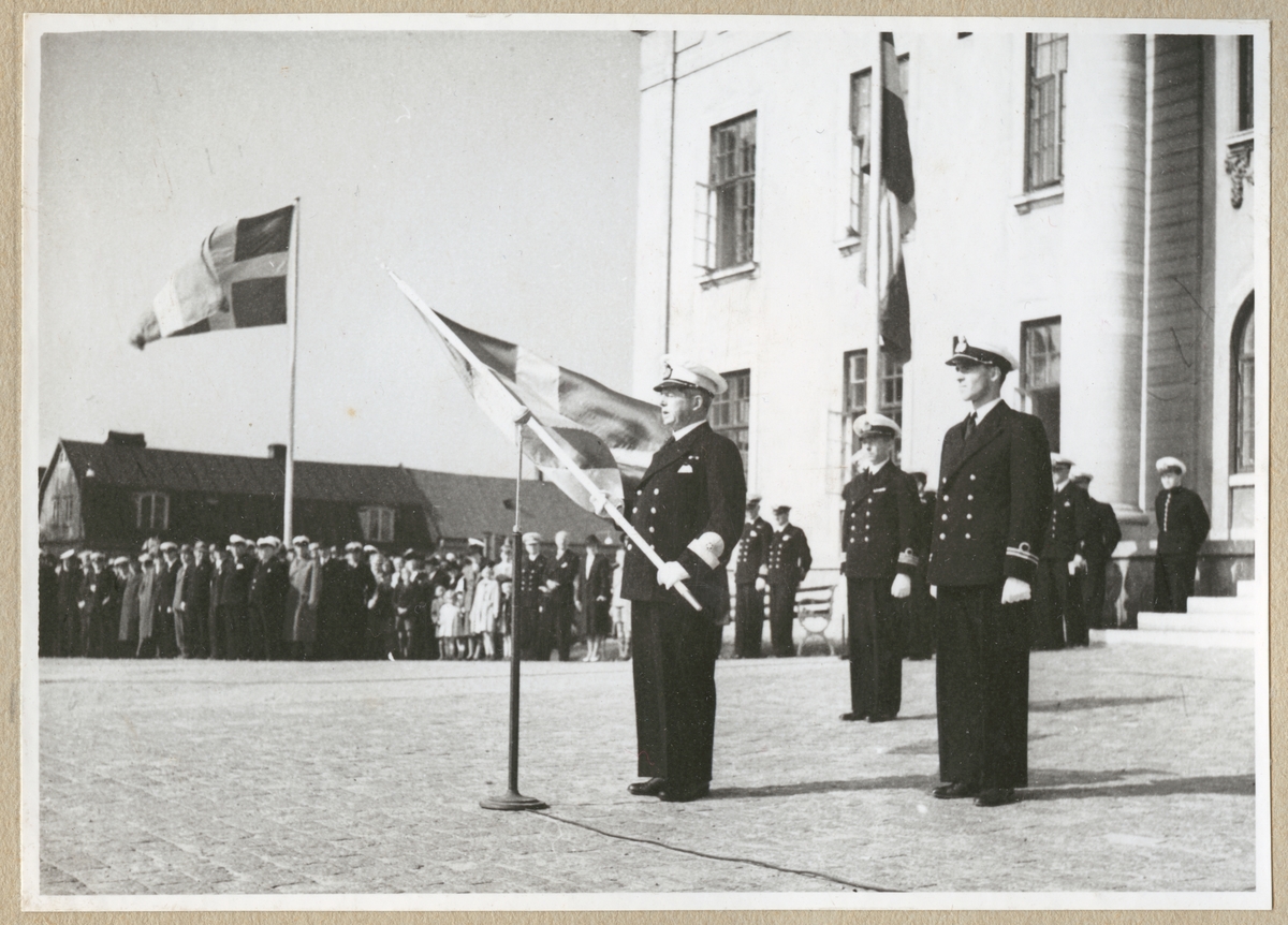 Konteramiral Ehrensvärd står på stortorget framför rådhuset i Karlskrona framför en mikrofon. I händerna håller han en stor Sverigeflagga. Bakom honom står en löjtnant och en kommendörkapten. I bakgrunden syns en folkmassa med blandade militärer och civila, samt fler flaggor.