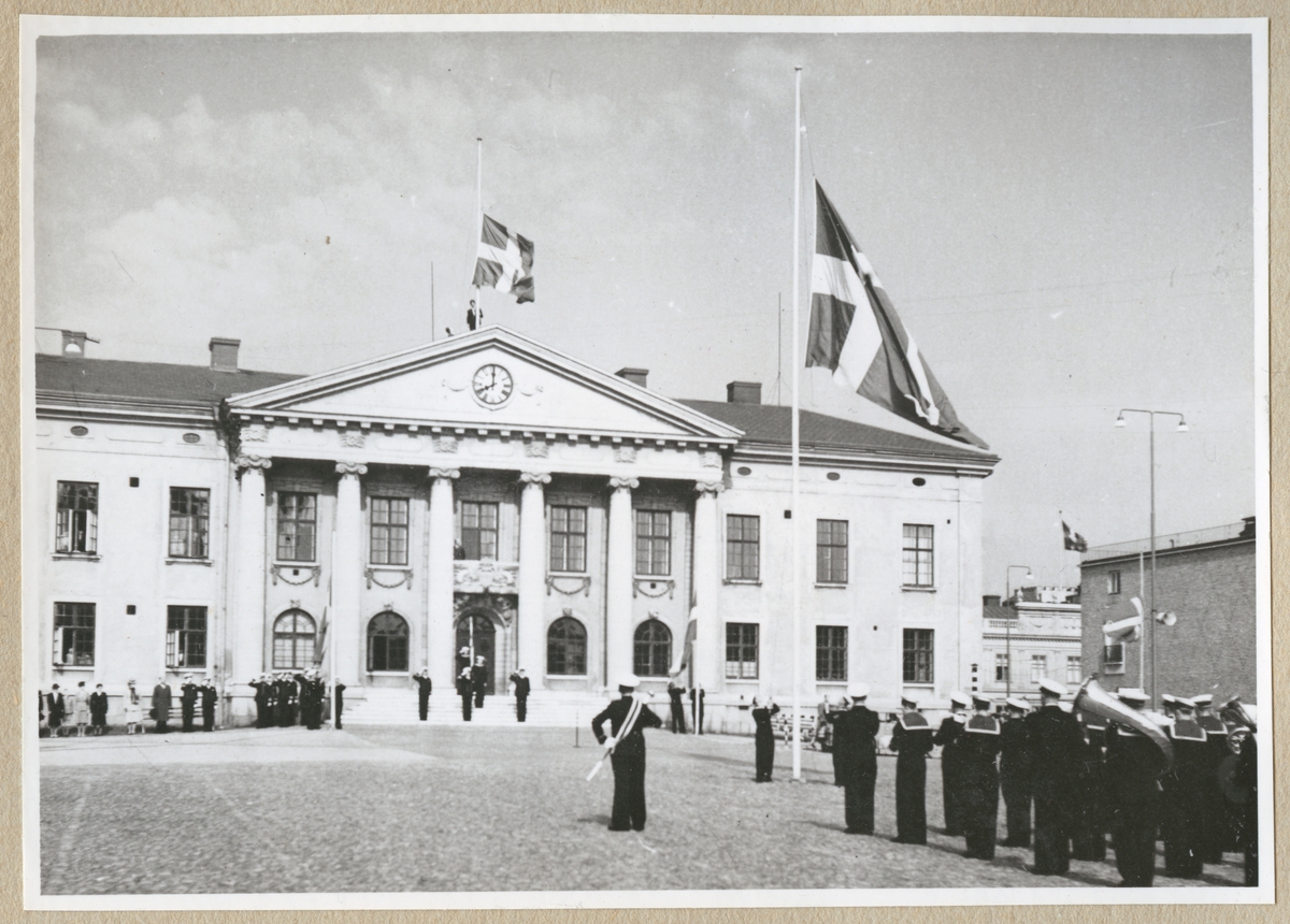 Marinens musikkår står på Stortorget i Karlskrona framför rådhuset och spelar medan fyra svenska flaggor, tre på torget och en på rådhuset, antingen hissas eller halas. På rådhusets trappa står ett par militärer och gör honör. Utanför rådhuset står några civila åskådare.