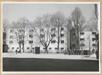 Uniformsklädda militärer står på en grusplan utanför Varvsgatan 3 i Karlskrona. Byggnaden består av två stramt utformade byggnadskroppar med putsade ljusa fasader. Byggnaden täcks något av en trädrad.