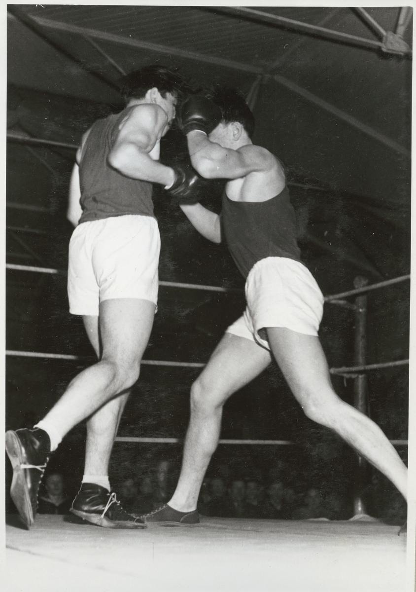 Två män står i en boxningsring och boxas. På golvet utanför ringen syns åskådare.