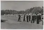 Uniformsklädda flottister och marinens musikkår med varsina svenska flaggor står på en gräsklädd yta. I bakgrunden syns grönskande träd.