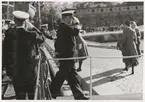 En konteramiral går av ett fartyg, förtöjt vid Kungsbron, via en landgång samtidigt som han gör honör. Ombord på fartyget syns ytterligare uniformsklädda militärer. På kungsbron står ett par kvinnor. I bakgrunden skymtar bastion Aurora och en folksamling.