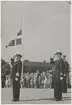 Konteramiral Gösta Ehrensvärd gör honör tillsammans med en kapten och en flottist medan en svensk flagga hissas. Alla tre är klädda i uniform och mössa. I bakgrunden syns ett plåtlagt tak och en folksamling bestående av barn och vuxna.