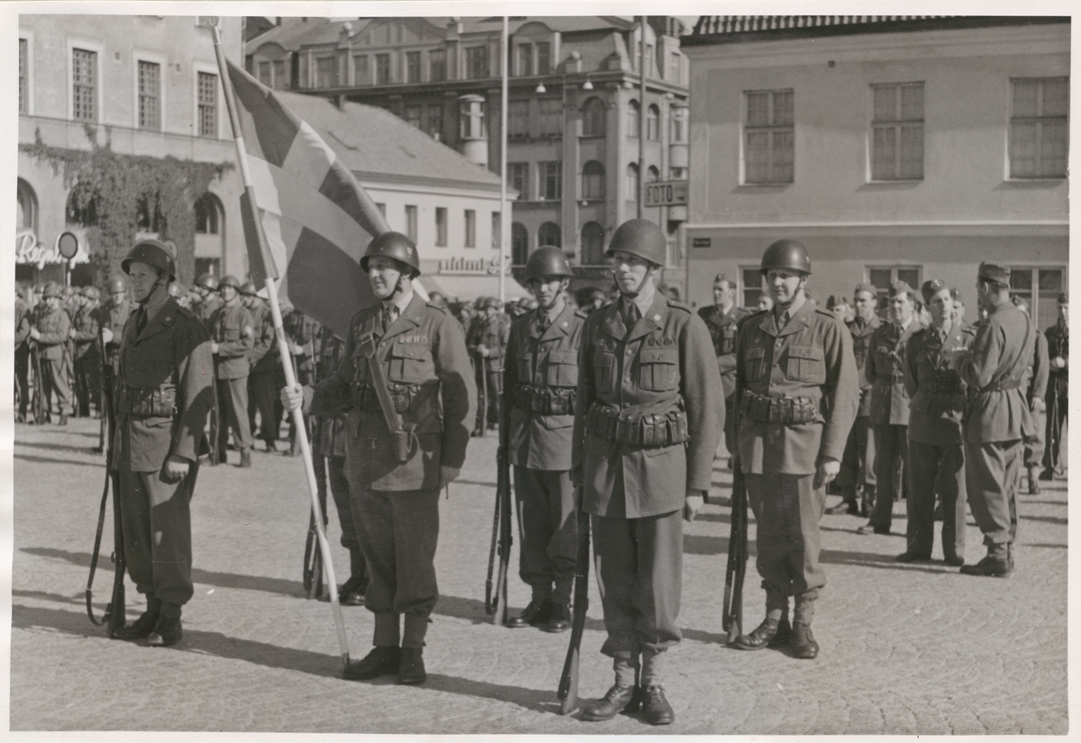 Uniformsklädda militärer från armén, utrustade med skjutvapen och hjälm alternativt mössa, står uppställda vid stortorget och Klaipedaplatsen i Karlskrona. En av militärerna håller i en svensk flagga. I bakgrunden syns omgivande bebyggelse omkring torget, Klaipedaplatsen och Ronnebygatan.