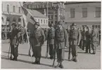 Uniformsklädda militärer från armén, utrustade med skjutvapen och hjälm alternativt mössa, står uppställda vid stortorget och Klaipedaplatsen i Karlskrona. En av militärerna håller i en svensk flagga. I bakgrunden syns omgivande bebyggelse omkring torget, Klaipedaplatsen och Ronnebygatan.