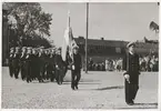 Flottister går på marsch på stortorget ledda av en fänrik, som håller en Svensk flagga, samt en kapten. I bakgrunden syns civila åskådare och byggnadstak.