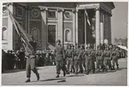 Flottister på marsch genom stortorget. Framför dem går en soldat med en svensk flagga. I bakgrunden syns civila åskådare uppradade framför Trefaldighetskyrkan.