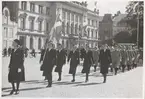 Kvinnor från lottakåren marscherar på stortorget i Karlskrona. Lottan näst längst fram håller i en svensk flagga. Samtliga lottor är klädda i uniform. De som går först har dock mörkare uniformer än resterande lottor. I bakgrunden syns Karlskrona stadshotell med sin rikt utsmyckade fasad.