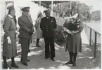 Konteramiral Gösta Ehrensvärd står tillsammans med lottor samt militärer från marinen och armén vid en läktare på en idrottsanläggnig, troligen Västra mark. Samtliga har uniform på sig. En av lottorna håller en blomsterbukett i famnen. Bakom dem står en yngre kvinna.