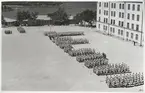 Uniformsklädda lottor står uppställda på Gräsviks övningsfält. Övningsfältet avgränsas i norr av den norra kasernen och i väst av rotundan.