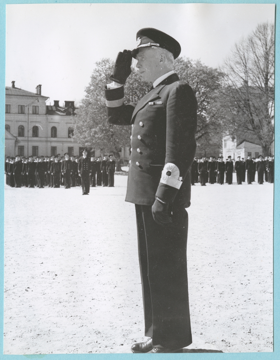 Viceamiral Strömbäck står på bataljon Sparres kaserngård och gör honör. Bakom honom syns uppställda militärer. Samtliga är klädda i uniform.