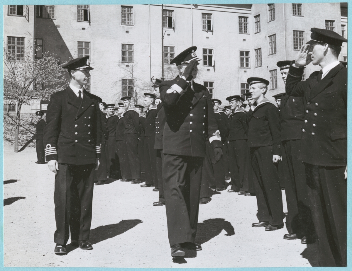 Viceamiral Strömbäck går tillsammans med en kommendörkapten av första graden och gör honör mot flottister uppställda på bataljon Sparrens kasernsgård. Samtliga är klädda i uniform. I bakgrunden syns kasern Sparre med putsad fasad.