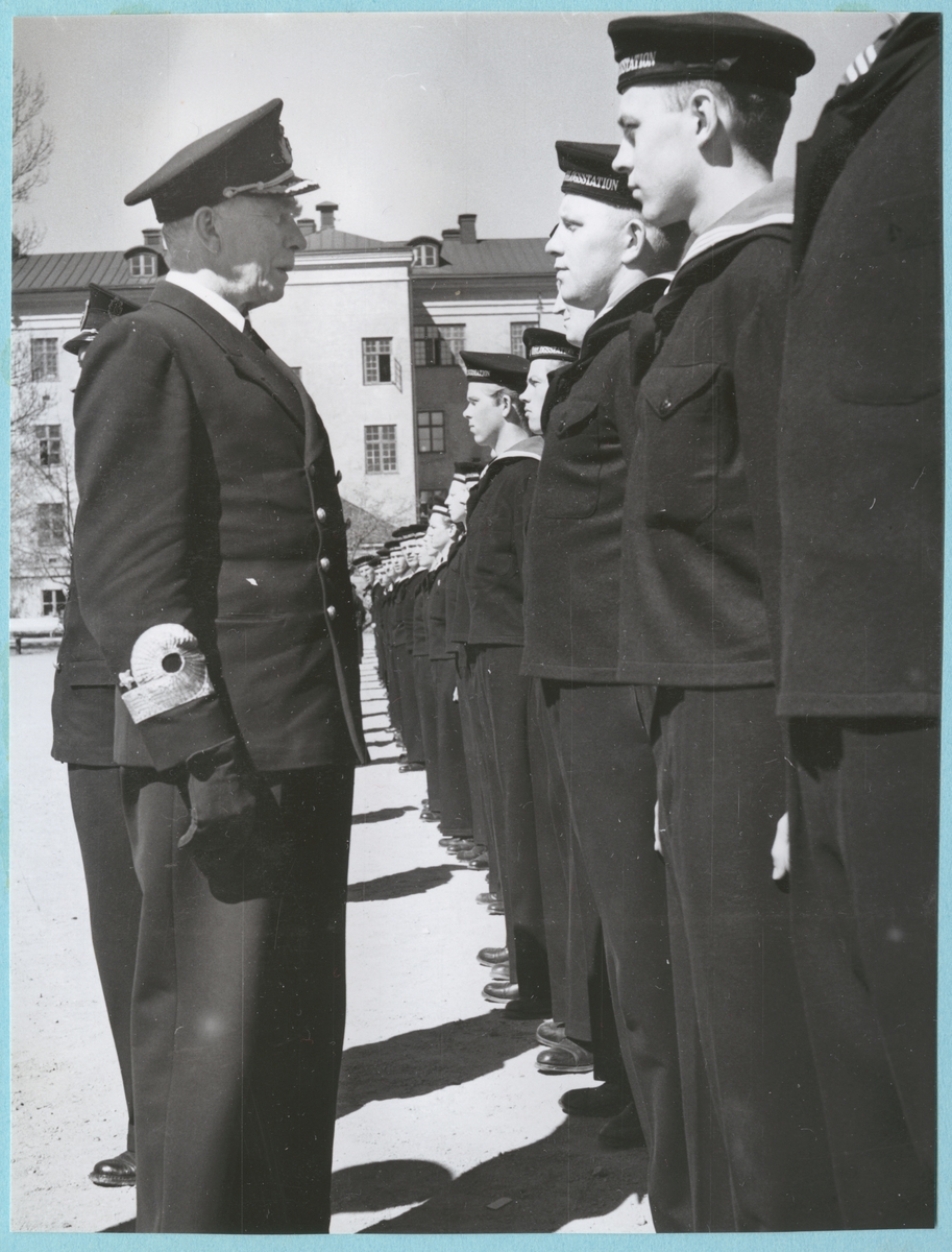Viceamiral Strömbäck står framför en rad med uppställda flottister på bataljon Sparres kaserngård. Samtliga är klädda i uniform. I bakgrunden syns kasern Sparre.