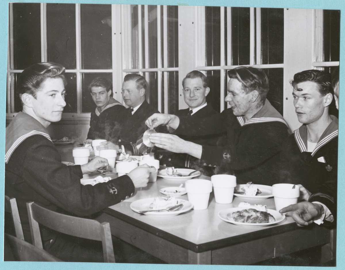 Uniformsklädda flottister från pluton 21 sitter runt ett dukat bord och äter fika och dricker kaffe. Med vid bordet sitter även två militärer av högre grad. Bilden är daterad till 25-2-1952.