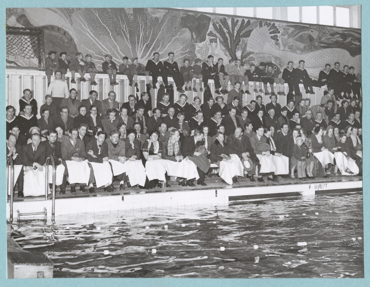 Flottister och civila åskådare sitter på läktaren i simhallen på Bataljon Sparre. Framför dem ligger simbassängen. Fotot är daterat till 15-3-52.