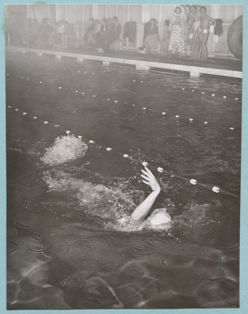 En simmare tar ett simtag på rygg i vattnet i en bana i en simbassäng. Längs kanterna på land står barn i simkläder samt vuxna människor i civila kläder som sitter på bänkarna. Fotot är daterat till 15-3-52