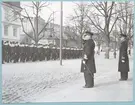 Uniformsklädda flottister marscherar längs med Drottninggatan. Vid sidan av gatan står konteramiral Samuelson och gör honör. Bredvid Samuelson står en liten bit bort en kommendörkapten av första graden. Fotot är daterat till 17-3-1952.