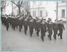 En grupp uniformsklädda flottister marscherar på Drottninggatan.  Längs med gatan syns militärer som tittar på marschen. Vid sidan om truppen syns byggnader till Bataljon Sparre.