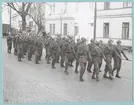 Uniformsklädda soldater, utrustade med hjälm och skjutvapen, marscherar längs med Drottninggatan. Den stenlagda gatan kantas av byggnader tillhörande bataljon Sparre.