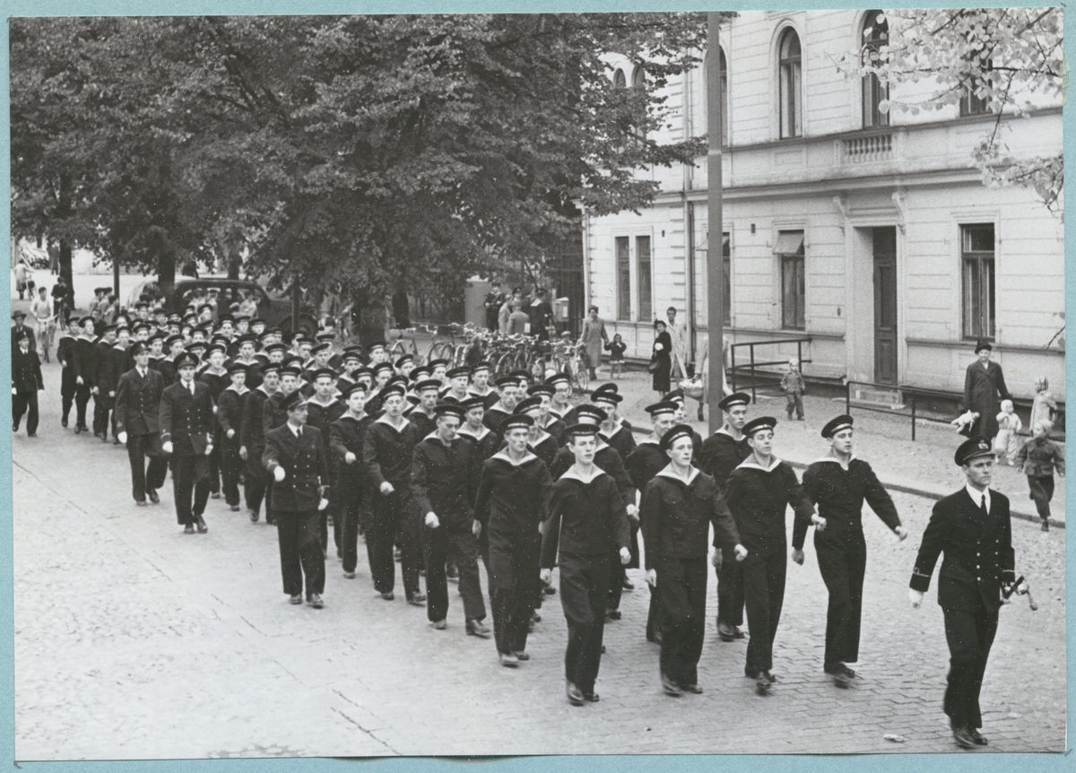 Bilder från Krigsmans erindran den 13/10 1951. Uniformsklädda flottister marscherar längs med den stenbelagda Drottninggatan. På gatan syns civila åskådare samt parkerade cyklar och en bil. Gatan avgränsas bl. a. bataljon Sparres kanslibyggnad.