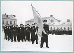 En grupp uniformsklädda flottister står uppställda på bataljon Sparres snötäckta kaserngård. Längst fram står en löjtnant med en flagga i ena handen. Kaserngården omgärdas av matinrättningen och exercishuset. Längs med exercishusets fasad står flera grupper av flottister uppställda.