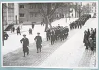 Uniformsklädda militärer marscherar längs med den snötäckta Drottninggatan. På gatan står både militärer och civila åskådare. Gatan kantas av bl.a. lägenhetshus med utskjutande balkonger. I bakgrunden skymtar också simhallen tillhörande bataljon Sparre.