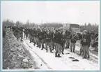 En trupp uniformerade soldater står på en snötäckt grusväg kantad av en stengärdsgård. I händerna håller de vapen. I bakgrunden syns en militär lastbil och trädtoppar i landskapet.