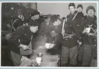 En militär står utomhus och serverar soppa i muggar under ett lätt snöfall. Runt omkring står flera militärer och köar, äter och pratar. Två av militärerna tittar in i kameran. Samtliga är klädda i uniform och mössa medan några har hjälm på huvudet.