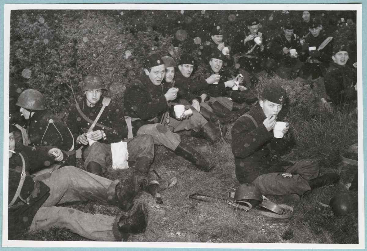 En grupp soldater i uniform med hjälmar eller mössor sitter på marken bland gräs och buskar och äter ur muggar. De ser in i kameran och vissa skrattar eller ler. I luften runt dem virvlar ett lätt snöfall.