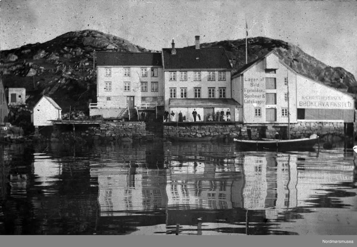 Fra venstre: skipper Strands vedbu, Strands bolig, bøkker Eriksens bolig, bøkkerverksted med skur foran, Eriksens brygge med torvskjul, Hjelkrembrygga (inntil 1904 Christie-brygga). På kaia og i båten sannsynligvis medlemmer av familiene Strand og Christoffersen. Se også  KMb-1984-000.0025. Fra Nordmøre museums fotosamlinger.