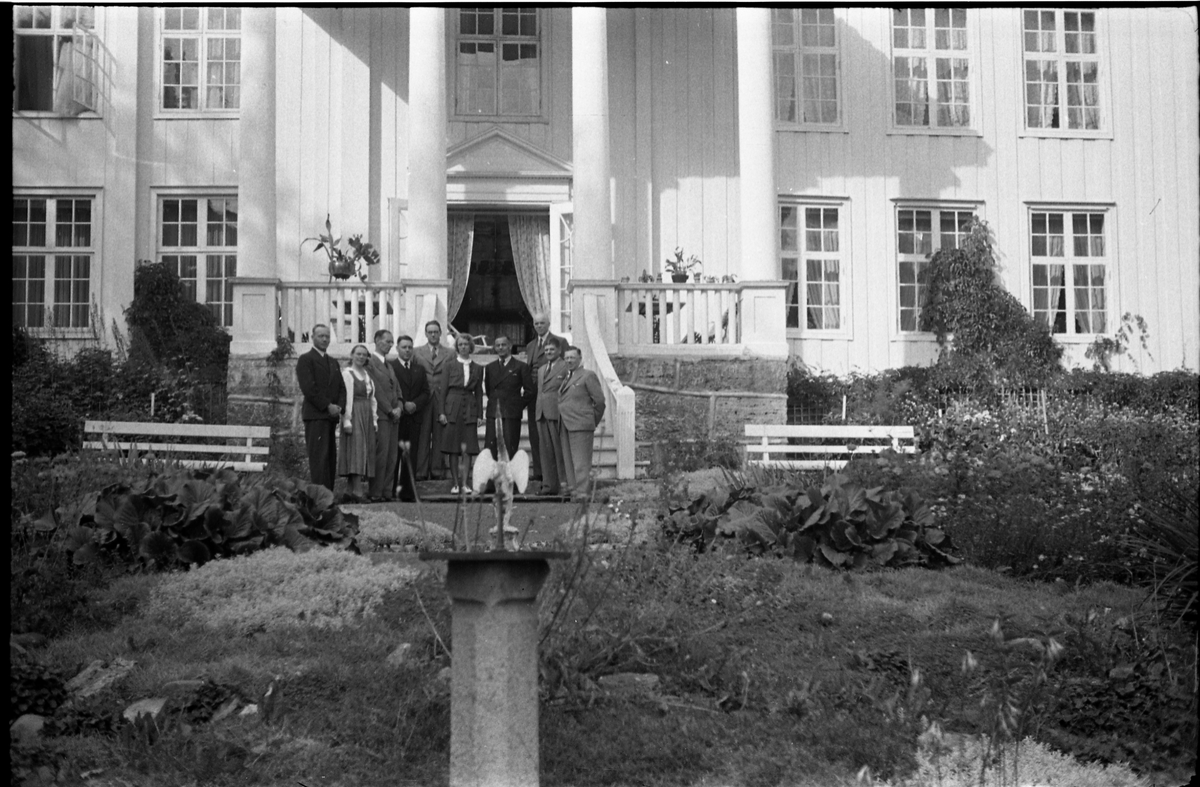 Noen personer, to kvinner og åtte menn, foran hovedbygningen på gården Rogneby, Østre Toten, august/september 1942.
Noen av personene er identifisert. Plasseringen refererer til bilde 1, fra venstre: Ukjent, Bergljot Holm, 2x ukjent, Sverre Wang, Eva Wang, ukjent, Adolf Holm (litt bak), Ragnar Gullichsen, ukjent.