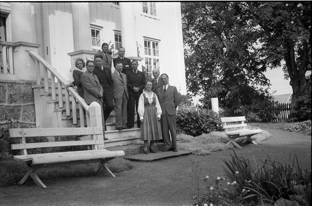 Noen personer, to kvinner og åtte menn, foran hovedbygningen på gården Rogneby, Østre Toten, august/september 1942.
Noen av personene er identifisert. Plasseringen refererer til bilde 1, fra venstre: Ukjent, Bergljot Holm, 2x ukjent, Sverre Wang, Eva Wang, ukjent, Adolf Holm (litt bak), Ragnar Gullichsen, ukjent.