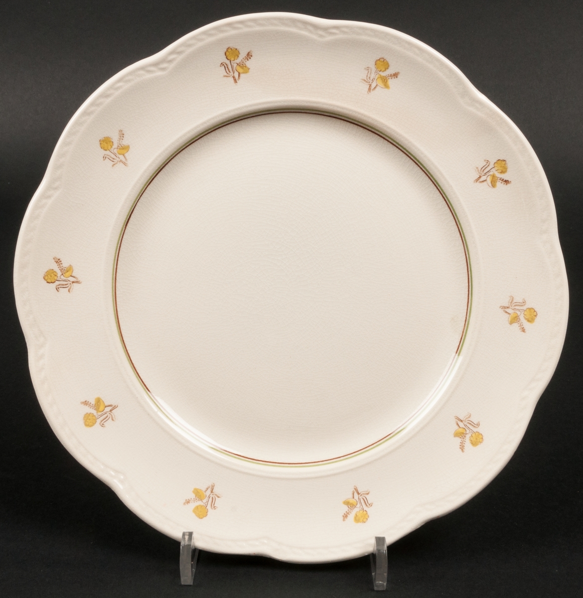 Flat mattallrik från Gefle Porslin AB. Dekor i form av gula blommor, brun och grön linje. Formgiven av Helmer Ringström. Tillverkad mellan åren 1945-1956.