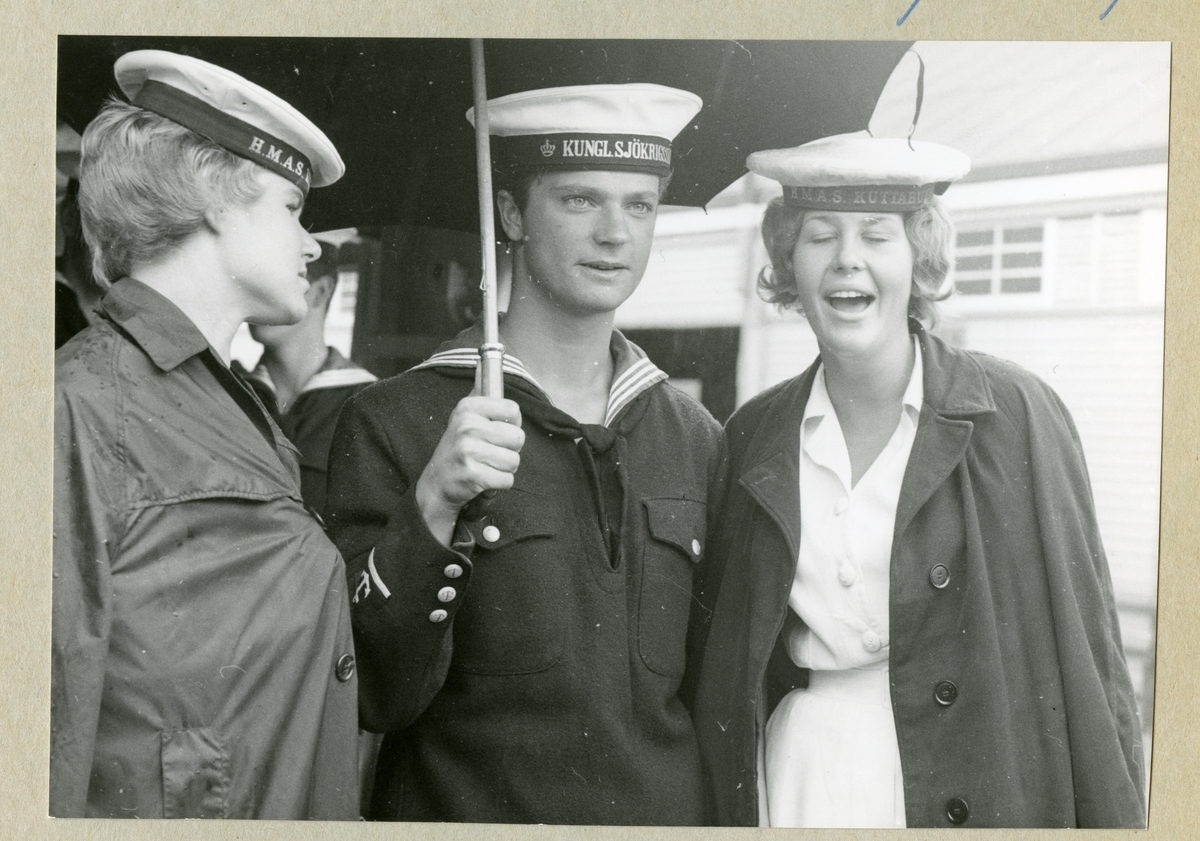 Bilden föreställer två kvinnor som står på varsin sida om kronprinsen Carl Gustaf, som är klädd i uniform och håller i ett paraply. De omgivande damerna är från HMAS Kuttaburra, en australisk marin utbildningsenhet. (HMAS = Her Majesty's Australian Ship).