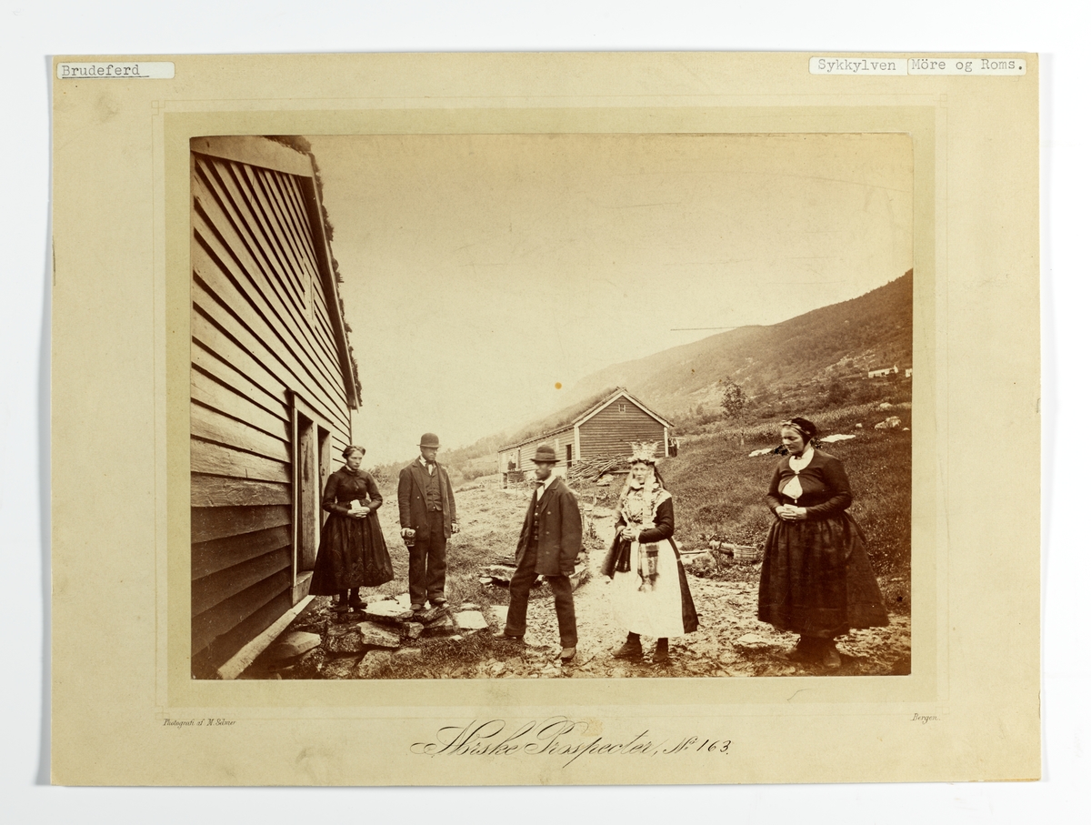 163. Brudeferd, Gjerdetunet, Straumgjerde, Sykkylven ca.1862. Brudepar, samt 3 personer utenfor hus på tun. Brudeparet er Elling P. Strømmegjerde og Louise Hansdtr. Fet. (Oppl. fra Sykkylven kommune, Møre og Romsdal. 1991). 
Inngår i serie "Norske Prospecter" (no.163) fot. av Marcus Selmer (1819-1900), Bergen.