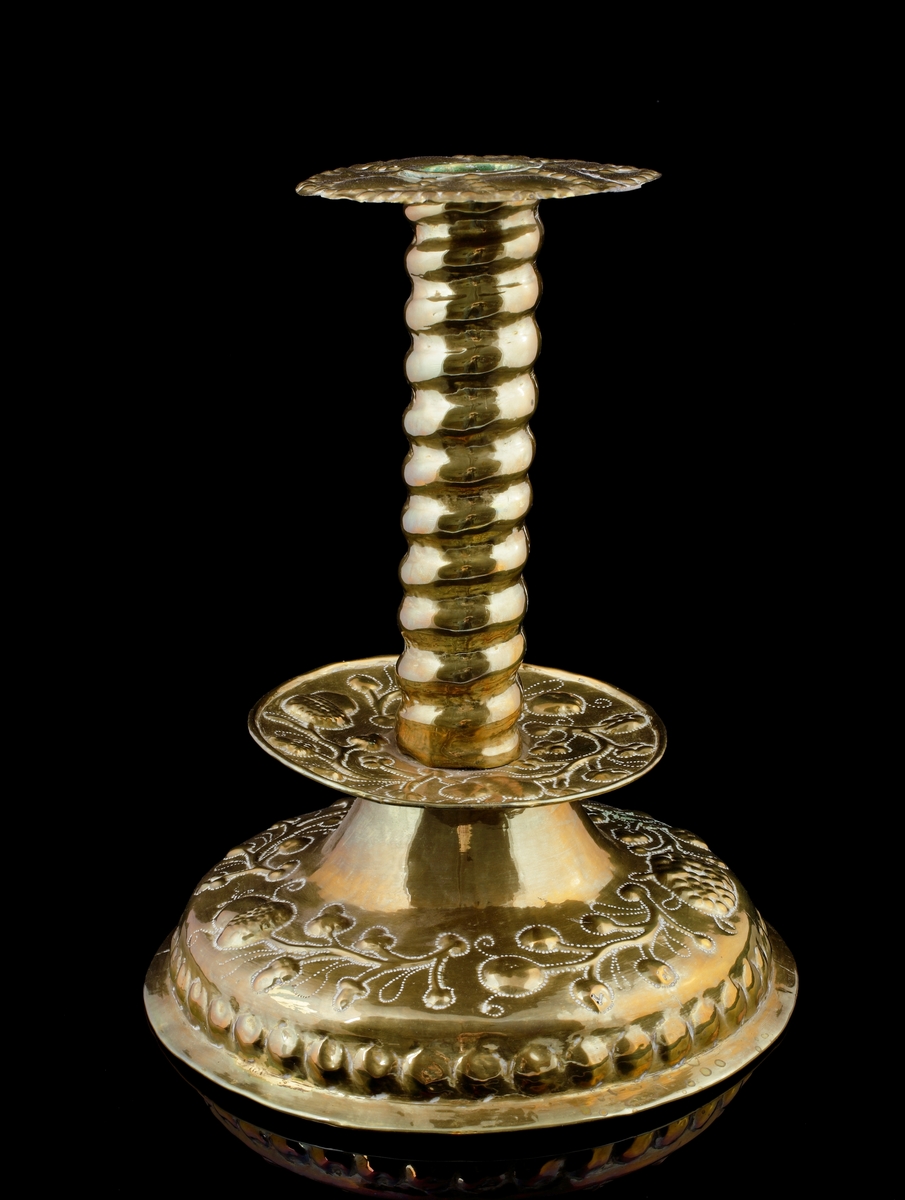 Barockljusstake av mässing, med rund fot och spiralvridet skaft. 
Foten är något kupad samt dekorerad med uthamrade växtornament.