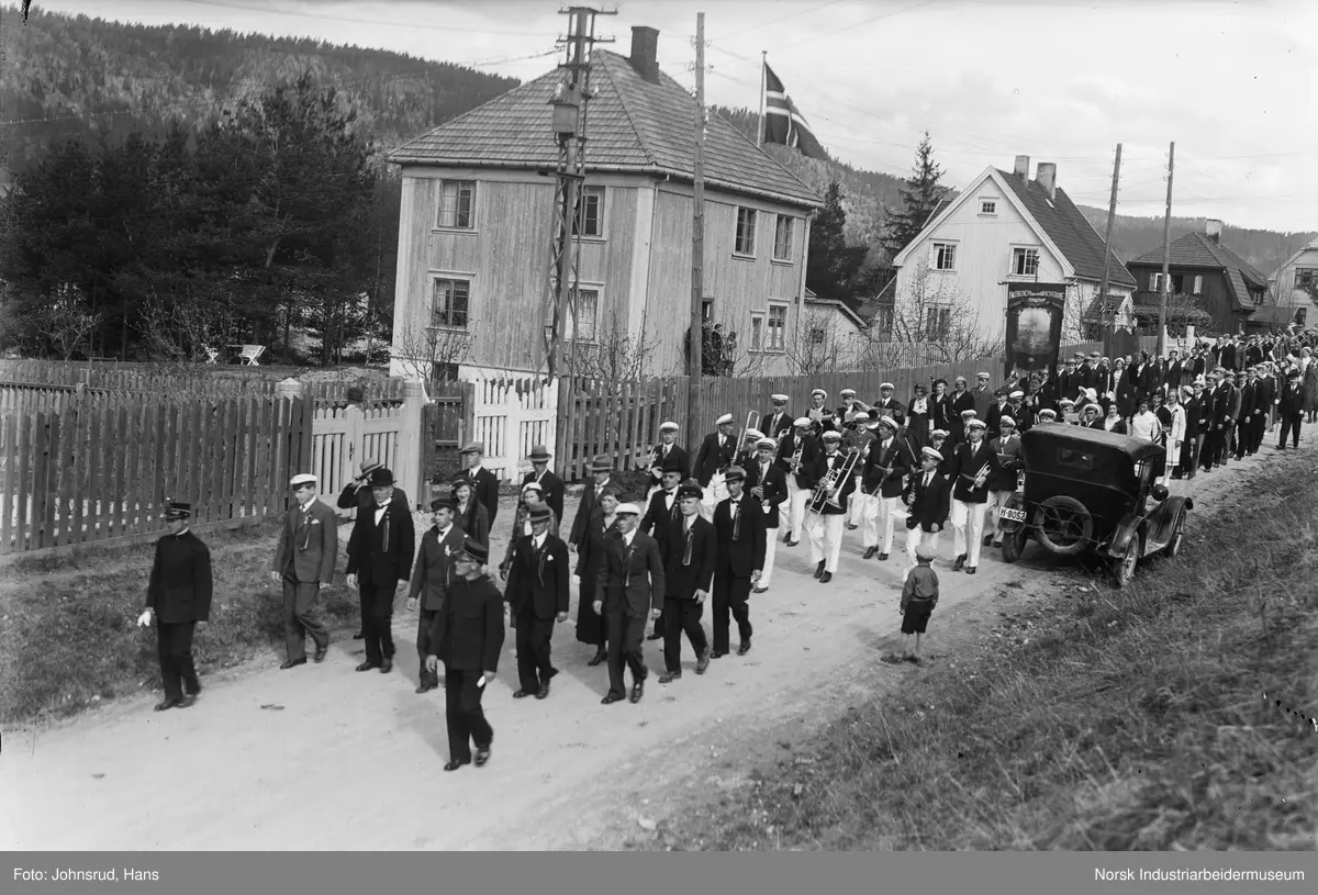 17. mai 1933. Russ med russelue og russestokk marsjer langs Roald Amundsens gate med skolefane. Politimenn og korps i front. Bil parkert langs veien. Flagg på flaggstang ved bolighus.
