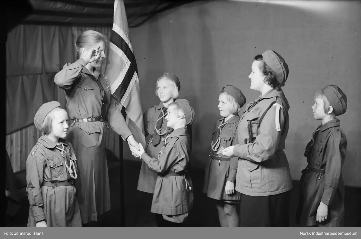 Gruppe speidere i uniform under speideropptagelse. Speidere hilser med speiderhilsen. Ragne Johnsrud holder det norske flagget.