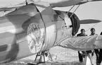 Flygplan Gloster Gladiator gul A, tillhörande F 19, Svenska frivilligkåren i Finland, märkt med 'Sammy Satana Perkele'. Vy av sidan, snett bakifrån. Två män står vid flygplansvingen.