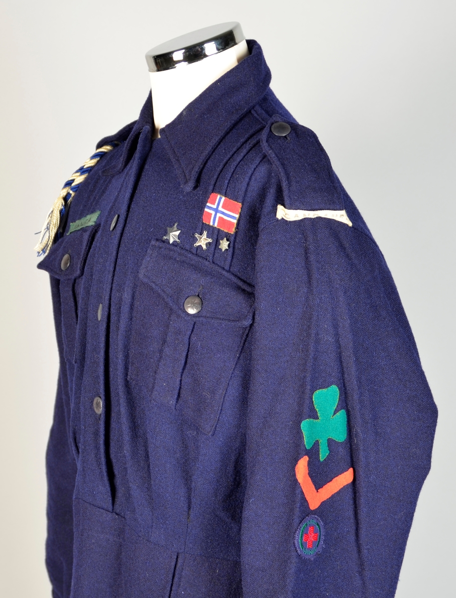 Speiderkjole i mørk blått ullstoff med grønt slips i bomull Skjørt og overdel er samansydd. Opning midt framme i overdel. Lukking med 4 knappar. 4 påsydde lommer framme, 2 på brystet, 2 på skjørtet. Klaff over lomma med knapp på lommane. Tre metallstjerner og eit norsk flagg er sydd på over venstre brystlomme. Over høgre brystlomme er sydd på band med "LINNEA". Blå og kvit silkesnor knytt inn på høgre skulderreim. Band med "SANDANE 1" sydd på venstre skulder. På venstre erme er det påsydd i filt: eit grønt trekløver, ein raud vinkel og eit rundt merke med ein raud kross inni og teksten: "GIRL GUIDES".