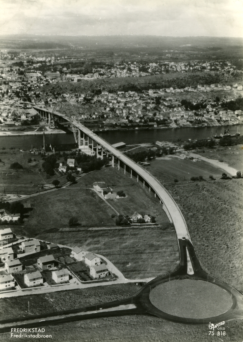 Fredrikstad. Vestsiden lensgt borte, Østsiden nærmest. Fredrikstadbroa. åpnet 18. august 1957. Frem til da hadde all trafikk mellom Vestsiden og Østsiden foregått med ferjer. På Østsiden sees Prestelandet og Gudeberg-området mot elva.  Det norske Cheviotspinneriet til høyre for broa. Broholdet i nedre bildekant var Fredrikstads første rundkjøring og ble tatt i bruk i forbindelse med broåpningen. Nabbetorpveien går under broa, r fra Prestelandet og nordover, frem til tettstedet Nabbetorp. utenfor høyre bildekant. - På Vestsiden ender broa på Kapellfjellet, Mot høyre i bildet Lahellemoen, Lahelle (ved elva) og Kiæråsen.  - Flyfoto: Harstad.