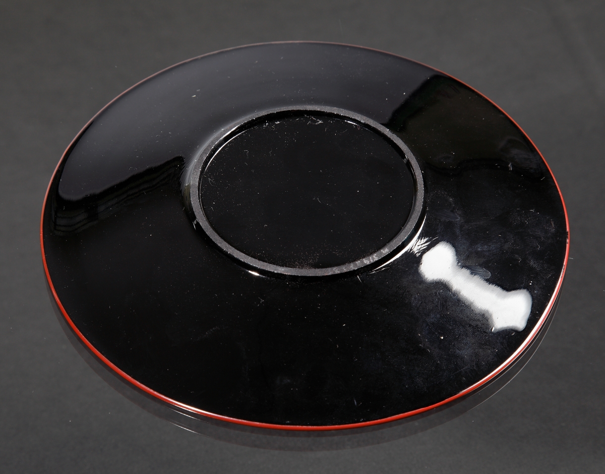 Assiett.
Teknik: Pressat, slipad bottenring, målad.
Färg/form: Svart degelglas, rund. Målad i rött, vitt och gult.
Dekor: Blommor likt blomman för dagen.
Målad av Karl Diessner (målning identifierad av Susann Diessner 2021-04-20).
Givarens mormor berättade att hennes morbror, Hans Teodorsson eller hans son George Hansson tillverkade föremålen. De arbetade på Eda innan de flyttade till Småland.
Givarens (Sivs) mormor fick föremålen av Hans eller George.