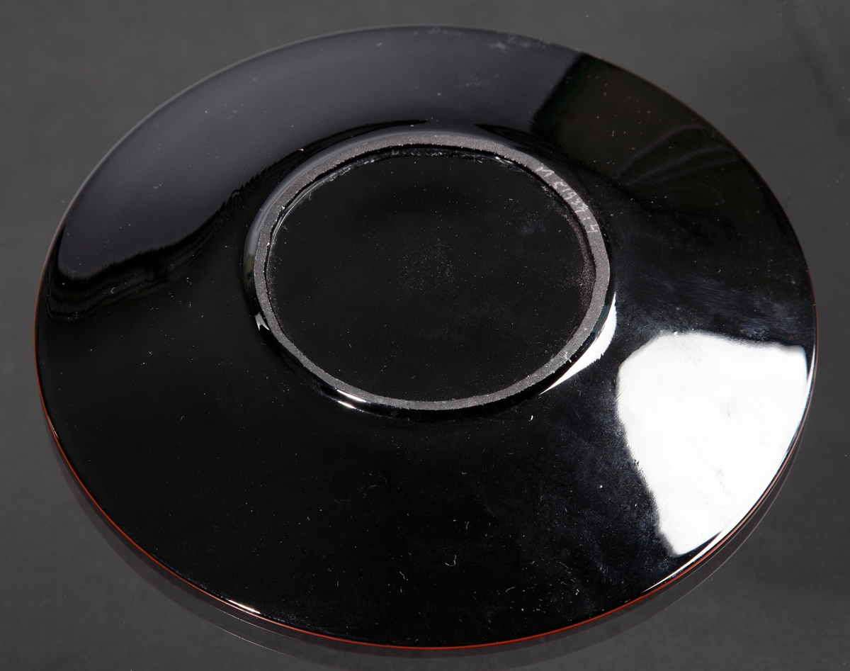 Assiett.
Teknik: Pressat, slipad bottenring, målad.
Färg/form: Svart degelglas, rund. Målad i rött, vitt och gult.
Dekor: Blommor likt blomman för dagen.
Målad av Karl Diessner (målning identifierad av Susann Diessner 2021-04-20).
Givarens mormor berättade att hennes morbror, Hans Teodorsson eller hans son George Hansson tillverkade föremålen. De arbetade på Eda innan de flyttade till Småland.
Givarens (Sivs) mormor fick föremålen av Hans eller George.