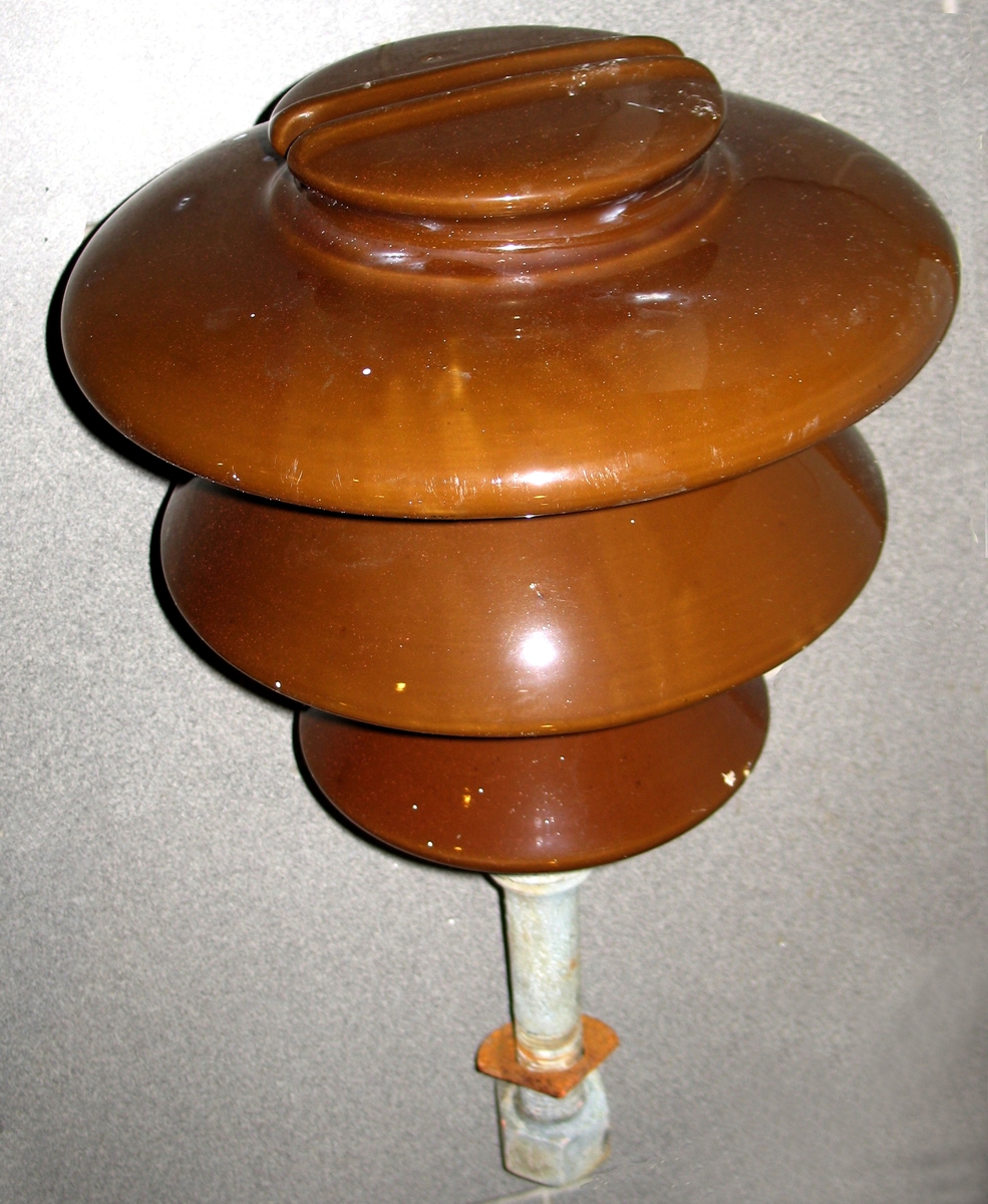 Isolator til bruk i kraftlinjer. Festebolt i utspart senterhull i isolatoren. Bolten er gjenget i enden, og det medfølger en skive og en mutter. Brun farge, det finnes også grønne av samme type. Typen isolator vises på forsidebilde i boken Kraft og børs, Arendals fossekompani 1896 - 1996.  På den ene er rester av kabel .