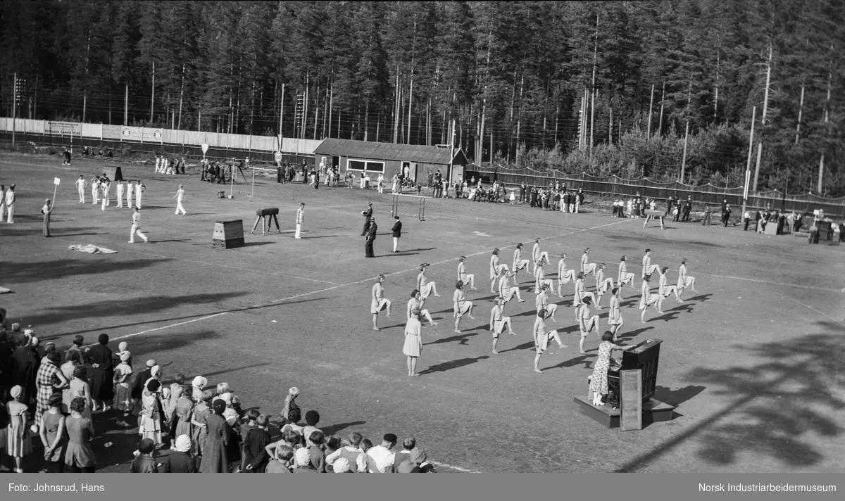 Turnstevne 1933. Turndamer har oppvisning, to kvinner ved piano stående på gresset. Menn som utøver turn på apparater ved siden. Tilskuere rundt stadion.