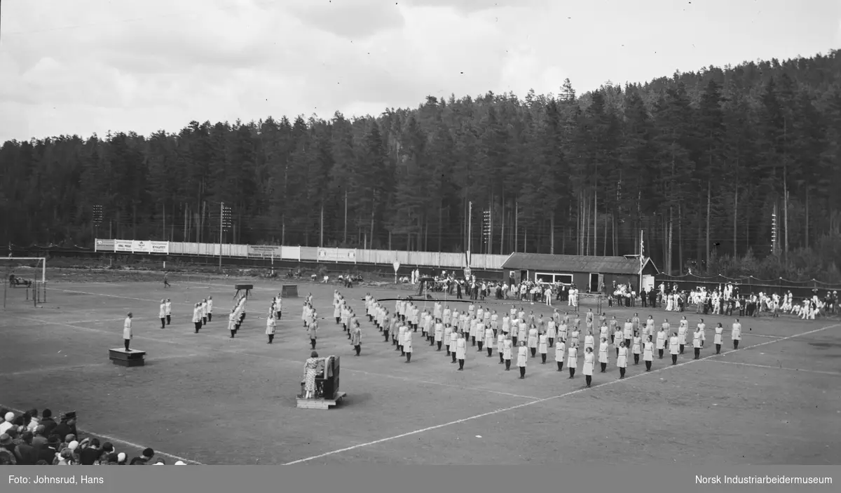 Turnstevne 1933. Turndamer har oppvisning, to kvinner ved piano stående på gresset. Tilskuere rundt stadion.