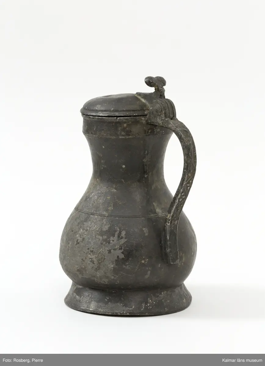 KLM 1886. Kanna, hankkärl. Av tenn. Hankkärl, bukig form, försedd med lock och handtag men utan näbb. Grepen ornerad på utsidan med bladranka som upptill avslutas med en djurliknande figur. Bottenmedalj i kannan, i form av korsfästelsemedalj. Spår efter förlorad lockmedalj, på lockets insida.