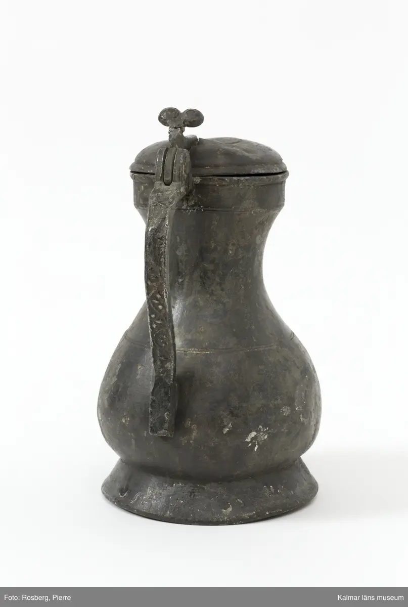 KLM 1886. Kanna, hankkärl. Av tenn. Hankkärl, bukig form, försedd med lock och handtag men utan näbb. Grepen ornerad på utsidan med bladranka som upptill avslutas med en djurliknande figur. Bottenmedalj i kannan, i form av korsfästelsemedalj. Spår efter förlorad lockmedalj, på lockets insida.